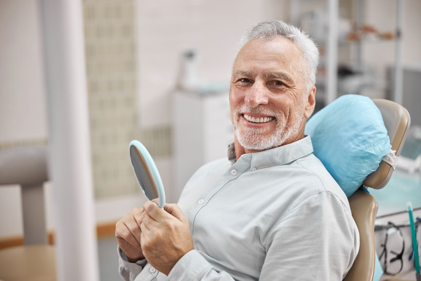 A Man Sitting On a Dental Chair Smiling While Holding a Mirror in His Hand
